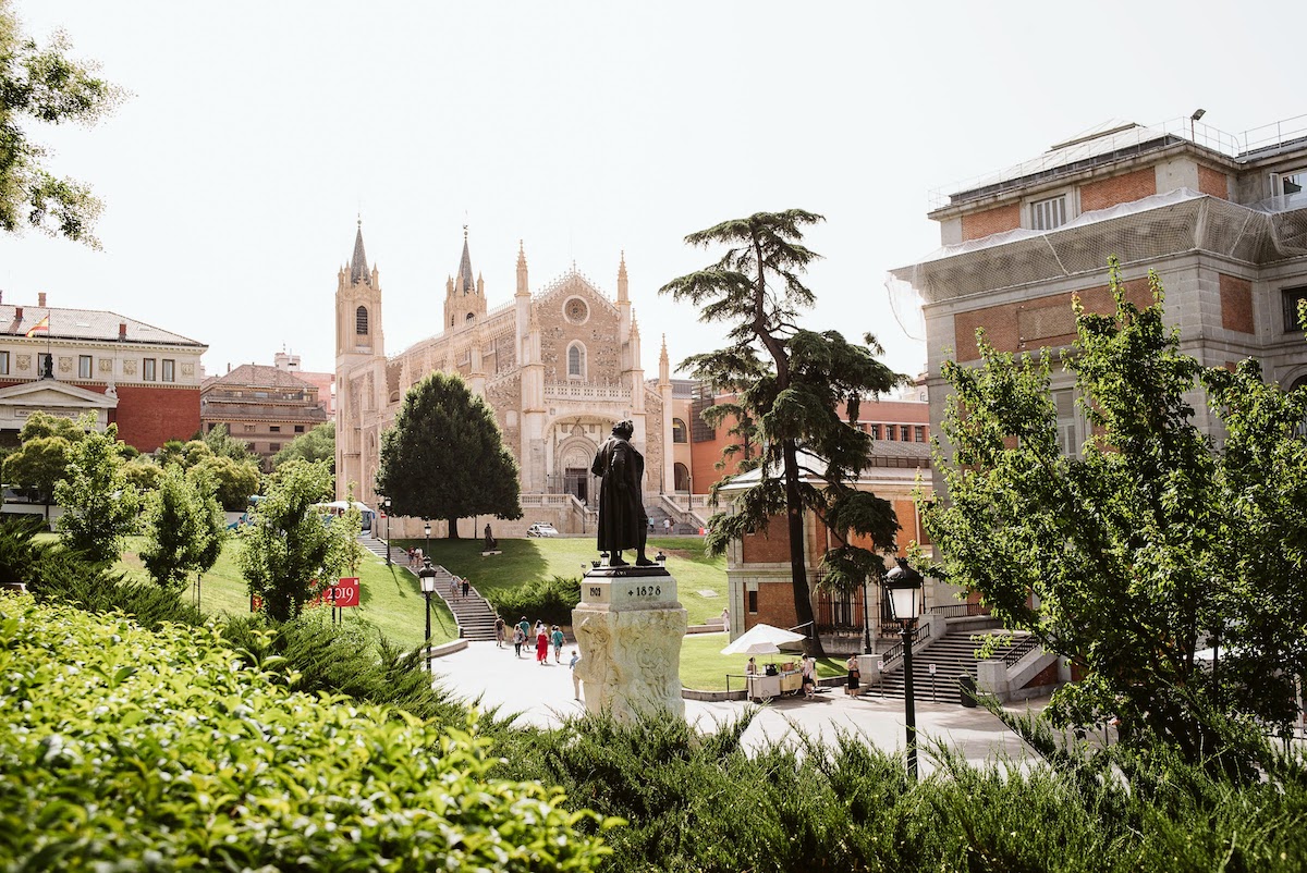 Escena de Madrid que representa una gran iglesia blanca, el Museo del Prado y una estatua de un hombre, todo rodeado de vegetación.