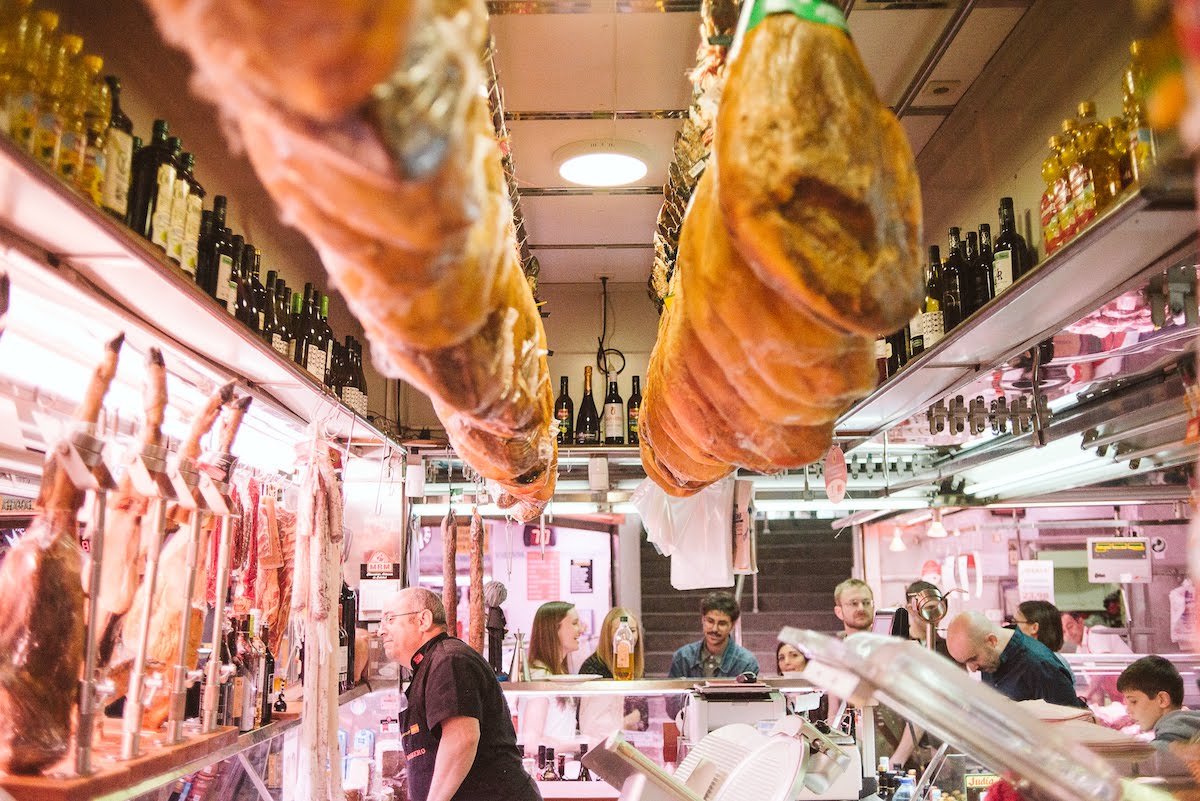 Un concurrido puesto de mercado en Madrid con patas de jamón curado colgando del techo.