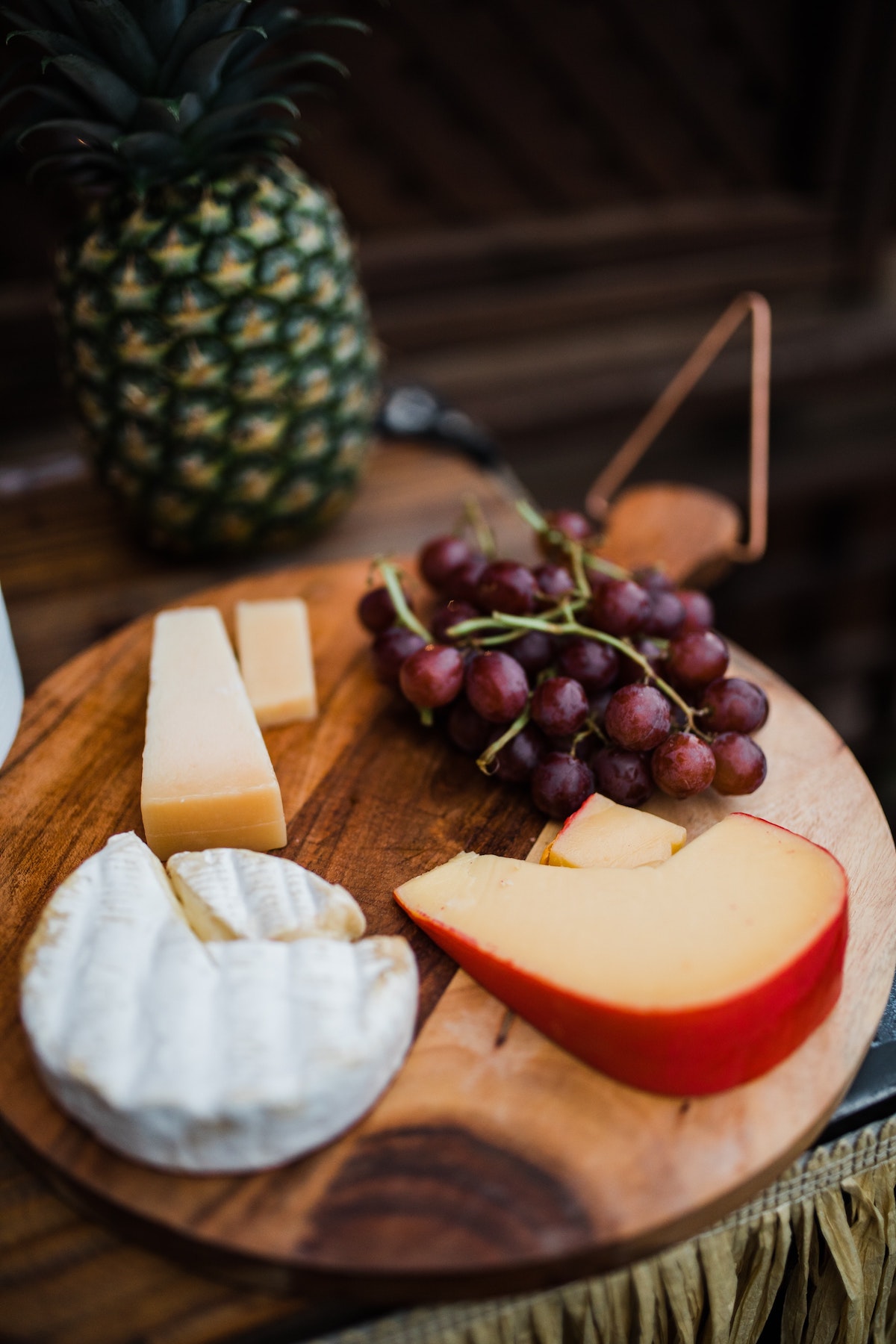 Tres tipos de queso en lonchas y un racimo de uvas rojas sobre una tabla redonda de madera con una piña visible al fondo.