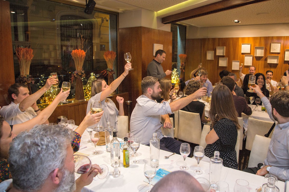 Un gran grupo de personas sentadas en las mesas de un restaurante levantan sus copas para brindar.