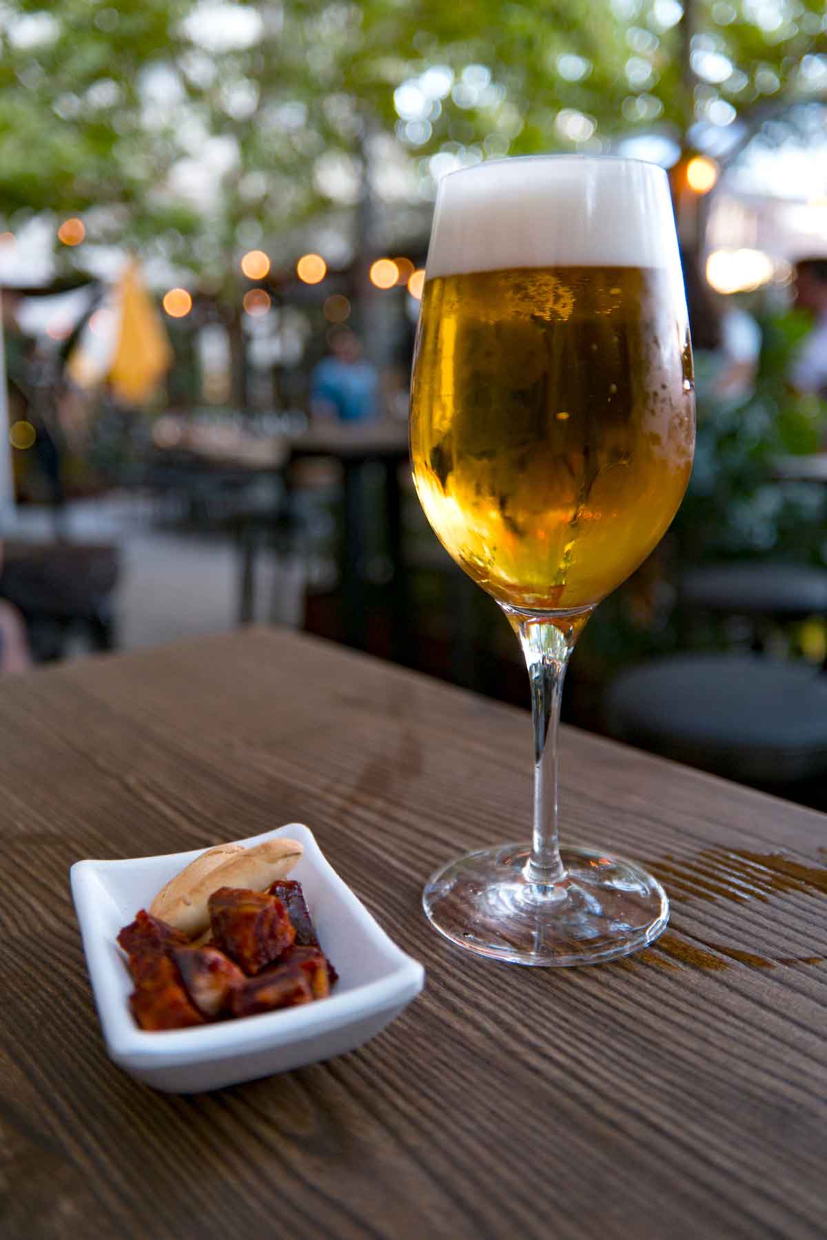Un vaso alto de cerveza junto a un pequeño plato blanco de trozos de chorizo y crujientes galletas de barra de pan sobre una mesa de madera en un espacio al aire libre.